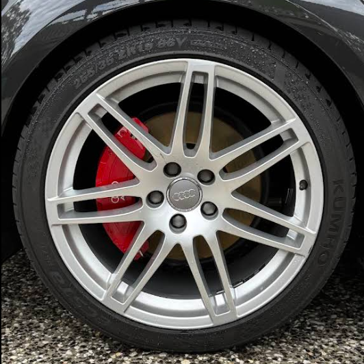 Close-up of a car wheel with a silver rim and red brake caliper, on a road with greenery in the background.