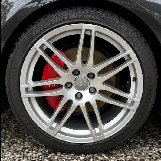 Close-up of a Audi allroad wheel with a silver rim and red brake caliper, on a road with greenery in the background.
